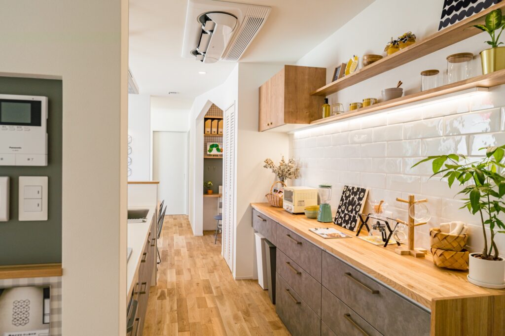 木目調カウンターとグレー収納がモダンなキッチン事例。白いタイル壁に間接照明、オープンシェルフにはおしゃれな小物や植物が並ぶ。