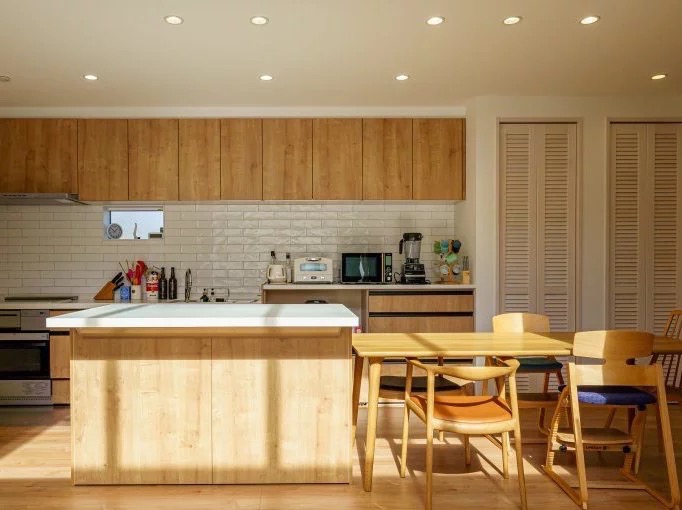 明るい光が差し込む、木目調で統一されたモダンなキッチンダイニング。白いタイル壁と木目調の吊り戸棚が特徴。