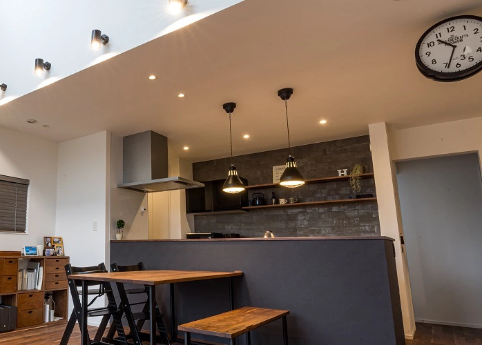 モダンなLDKのインテリアデザイン。ブラックのキッチンと木製のダイニングテーブルが並び、天井のダウンライトと2つのペンダントライトが空間を照らす。